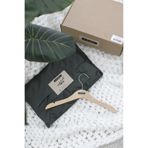 Ein schwarzes Paket mit einem Aufkleber, daneben ein Holzkleiderbügel auf einer weißen, gestrickten Decke und einer Pflanze im Hintergrund.