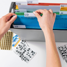 Hände sortieren farbige Ordner in einer Aufbewahrungsbox mit beschrifteten Trennblättern. Daneben liegen Aufkleber mit Buchstaben.