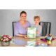 Eine Frau und ein Mädchen sitzen an einem Tisch, halten ein grünes Heft und lächeln. Auf dem Tisch liegen Schreibmaterialien und Blumen.