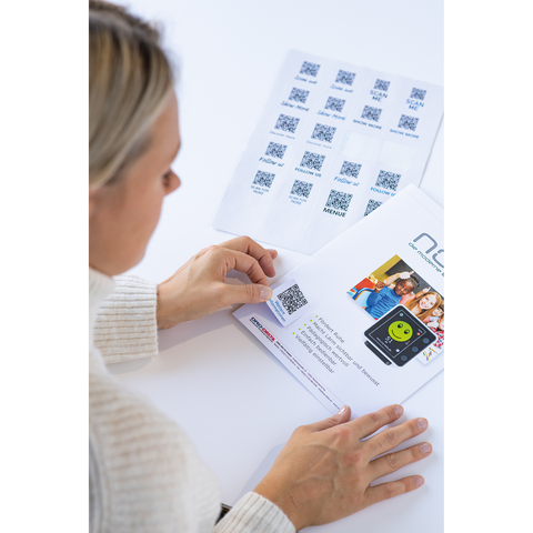 Eine Person scannt einen QR-Code auf einem Menüblatt, während ein weiteres Blatt mit mehreren QR-Codes im Hintergrund liegt.