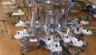Automatic labelling machine with multiple stations that applies labels to products. Control panel visible in the foreground.