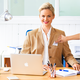 A woman with blonde hair is sitting at a desk, smiling and wearing a blue blazer. There is a laptop on the table.