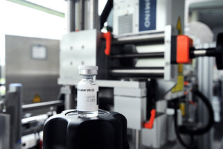 A vaccine vial is placed on a black lid, with machines for vaccine production visible in the background.
