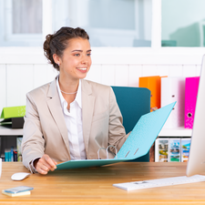 Eine Frau in einem hellen Anzug sitzt an einem Schreibtisch, hält eine Mappe und lächelt, während sie auf einen Computerbildschirm schaut.