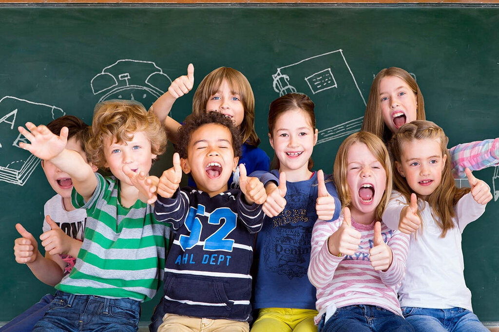 A group of children stands in front of a board, smiling and posing happily. Drawings of school supplies are visible in the background.