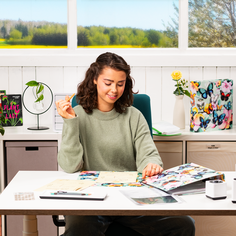 A young woman sits at a table, surrounded by notes and a laptop, while she gestures with one hand. In the background, there are windows overlooking a green landscape.