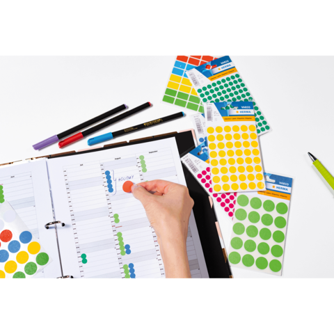 Eine Hand platziert einen roten Aufkleber auf einem Planer, umgeben von bunten Aufkleberbögen und Stiften.