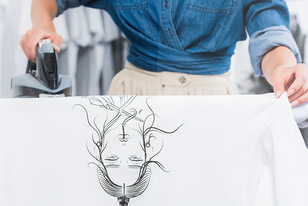 A person is ironing a white shirt with a graphic print while standing at an ironing board.