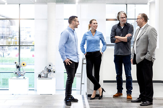Four people in a modern office are talking to each other, while in the background, an industrial building and a sprawling facility are visible.