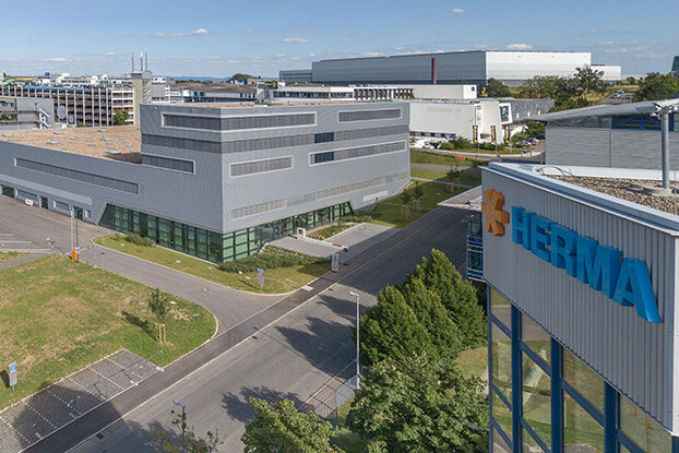Four people in a modern office are talking to each other, while in the background, an industrial building and a sprawling facility are visible.