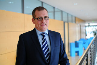 A man in a suit and glasses stands in a modern office hallway. He is smiling and wearing a striped shirt with a tie.