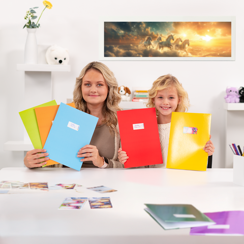 Two girls are sitting at a table holding colorful binders. On the table, there are photos, and in the background, there’s a picture of unicorns.