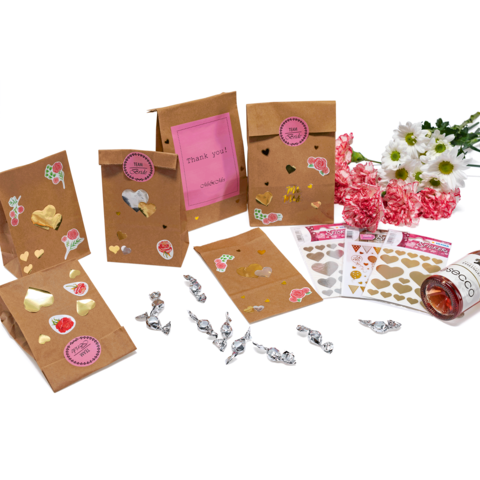 Geschenktüten aus Kraftpapier mit bunten Aufklebern, eine Flasche und Blumenarrangement, umgeben von Süßigkeiten und Dekorationen.