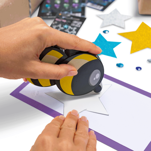 A hand uses a stamp to print a star on a sheet of paper, surrounded by colorful stars and decorations.