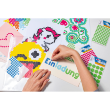 Eine Hand platziert bunte Klebepunkte auf ein Einladungskarten-Design, um ein kreatives Kunstwerk zu gestalten. Verschiedene Bastelmaterialien sind sichtbar.