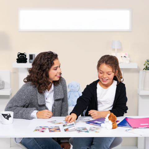 Two girls are sitting at a table, surrounded by photos and notes, as they talk and laugh together.