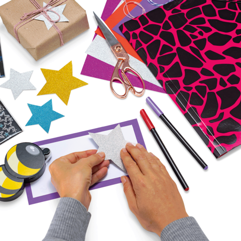 Hands sticking a glittering star onto colorful paper, surrounded by crafting materials like scissors, markers, and wrapping paper.