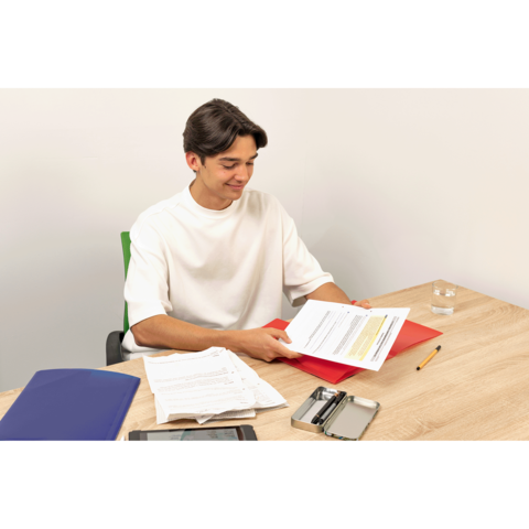Ein junger Mann sitzt an einem Tisch, hält Dokumente in der Hand und schaut auf seine Unterlagen. Auf dem Tisch liegen weitere Papiere und ein Smartphone.