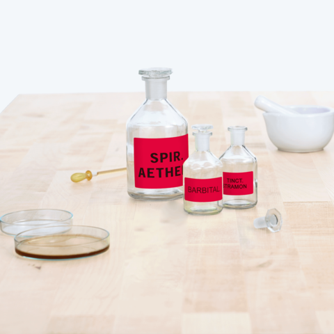 Flaschen mit roten Etiketten, beschriftet mit "Spir. Aether", "Barbital" und "Tinct. Tramon", stehen auf einem Tisch neben einem Mörser.