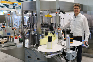 A man stands next to a modern bottling and labelling machine in a production hall, surrounded by bottles and machinery.