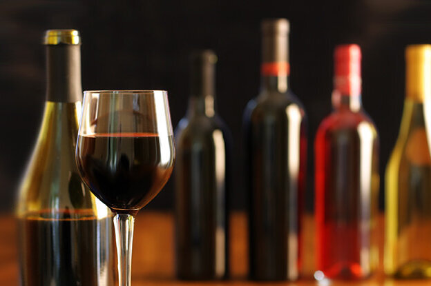 A glass of red wine stands in front of several wine bottles in different colours on a table.