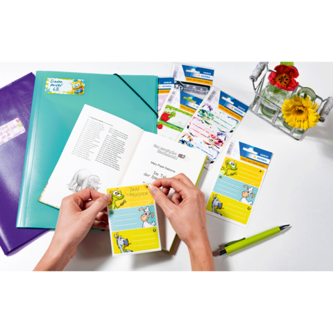 Hände halten einen bunten Notizzettel mit einem Cartoon-Motiv. Im Hintergrund sind ein Buch, Schreibwaren und eine Vase mit Blumen sichtbar.