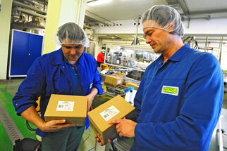 Two men in blue work jackets and hairnets are holding boxes in a production environment and examining them closely.