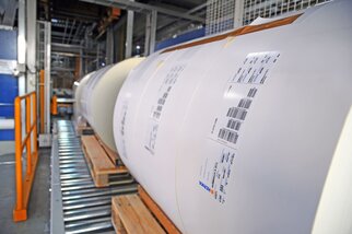 Rolls of paper on a conveyor system, each labelled with information and barcodes.