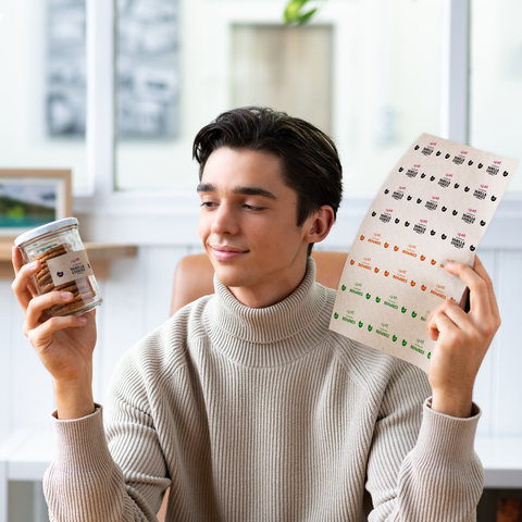 A young man is holding a glass of snacks in one hand and a label with various designs in the other.