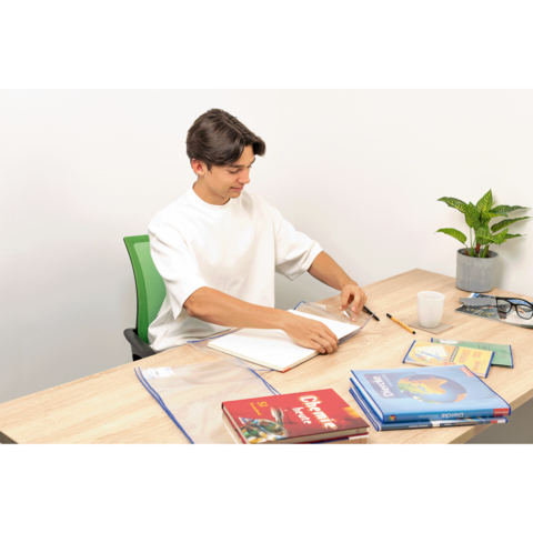 Ein junger Mann sitzt an einem Tisch, blättert in einem Notizbuch und hat Bücher sowie Schreibmaterialien vor sich. Eine Pflanze steht im Hintergrund.