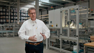 A man in a white shirt stands in a modern production environment with machines and shelves in the background.