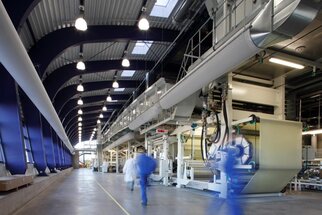 Modern production hall with machines and employees in work attire moving between the equipment. Bright lighting and high ceilings.