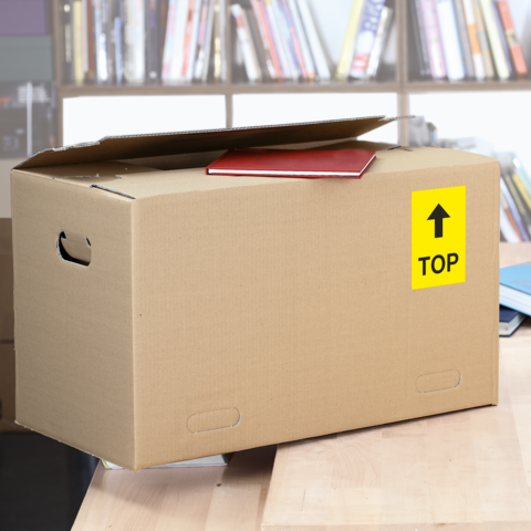 A cardboard box with a red notebook on top, labeled with a yellow 'TOP' sign, is sitting on a table in a library.