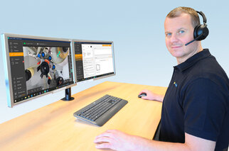 A man sits at a desk with two monitors displaying technical software and a camera view. He is wearing a headset.